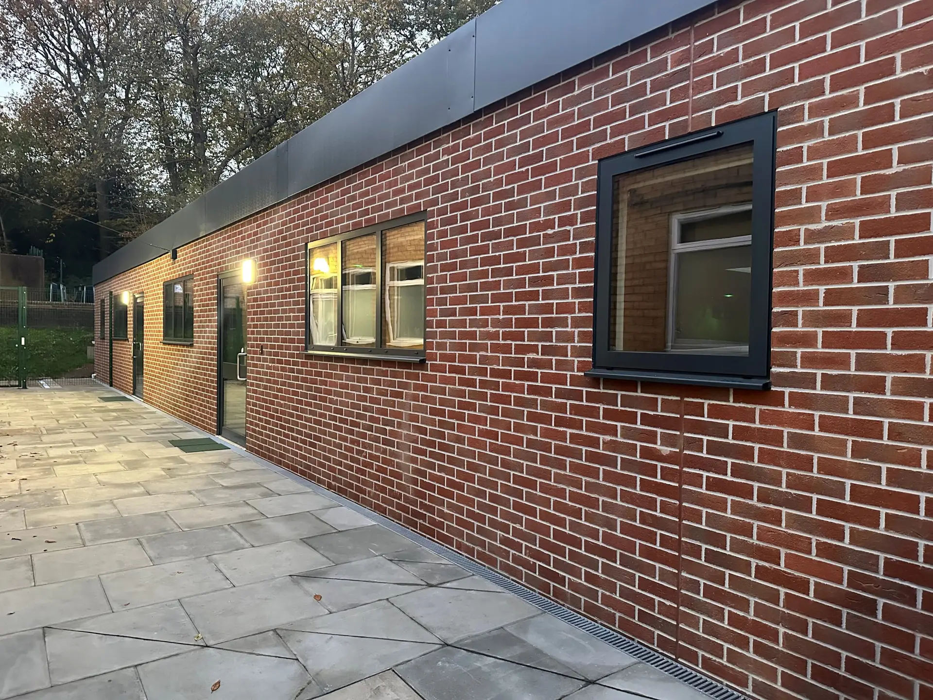 Side view of a modern red brick school building with large windows and a paved walkway, surrounded by trees.