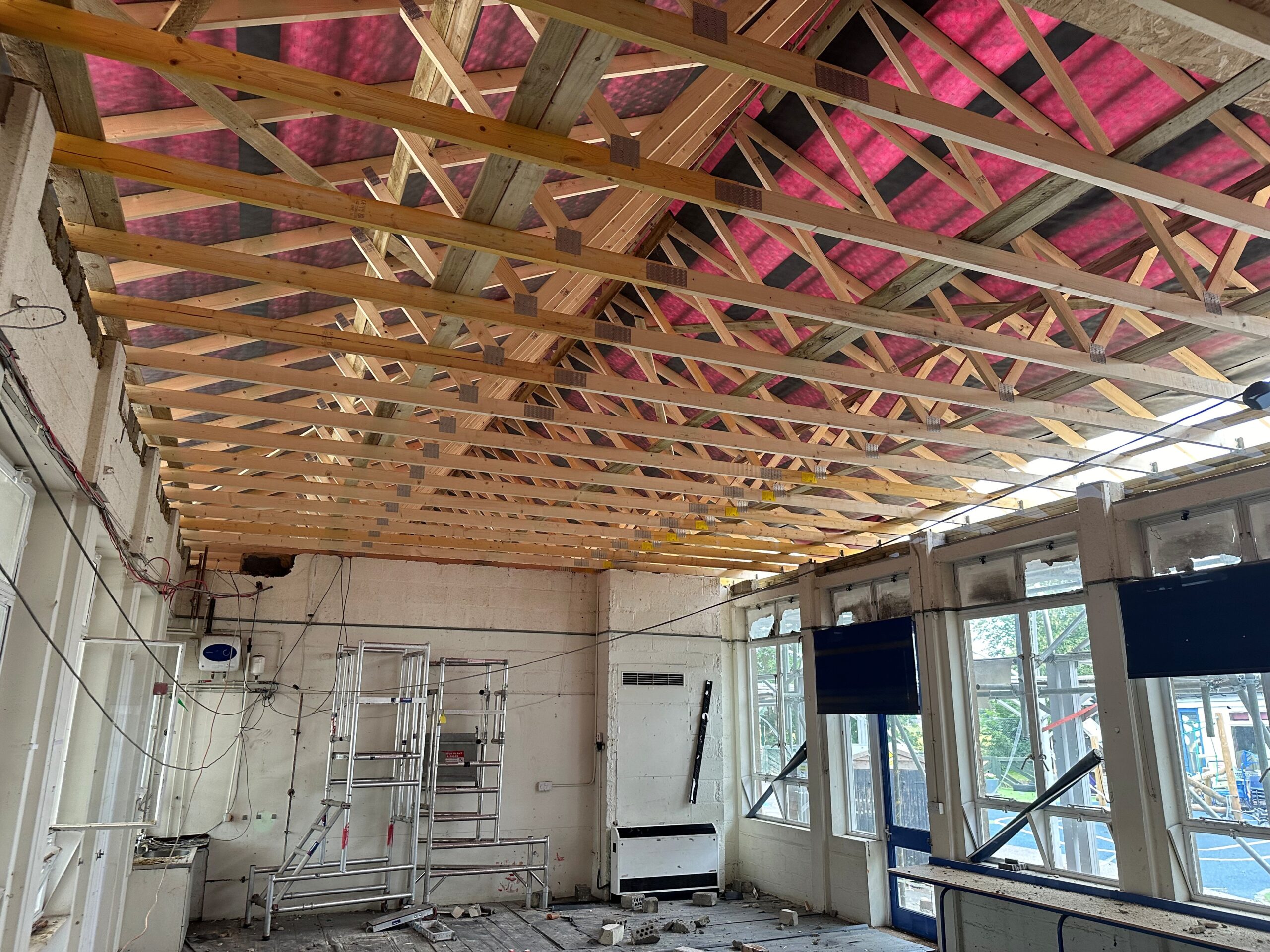 Interior classroom ceiling during construction, displaying new timber roof trusses and exposed services before insulation and ceiling installation.