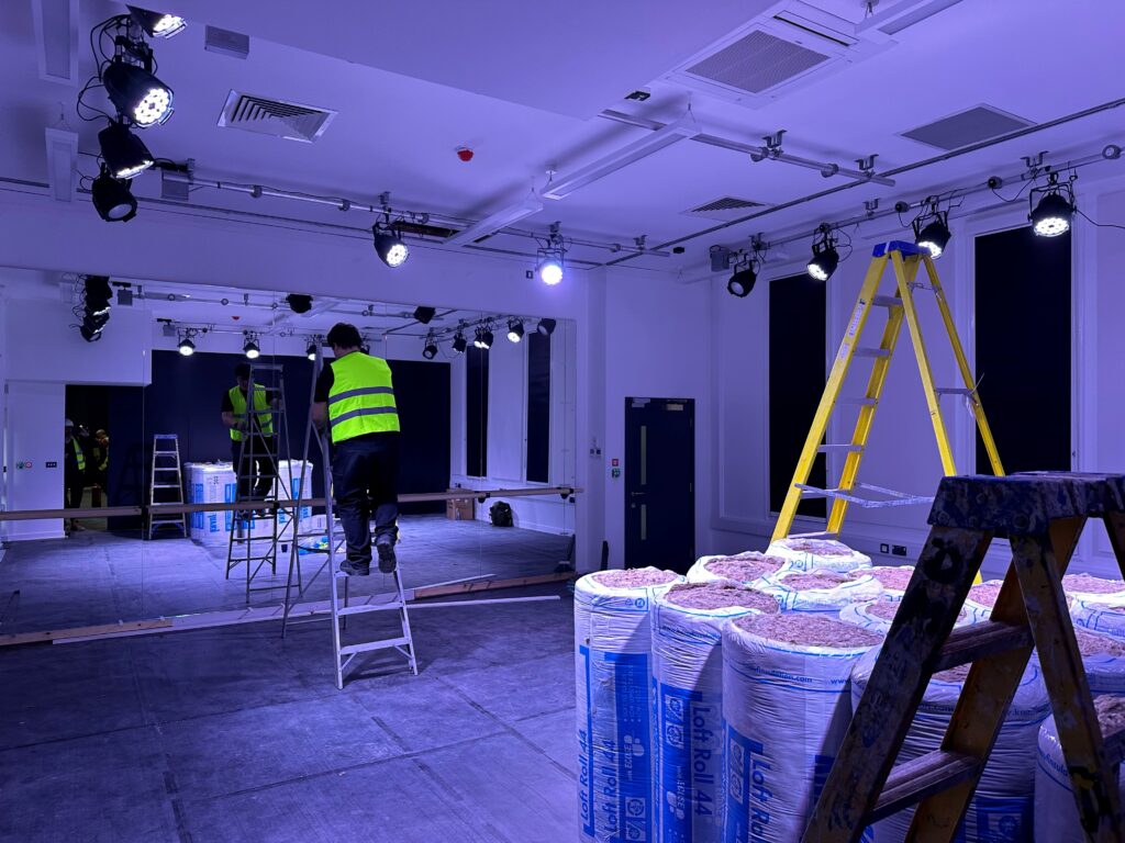 Construction workers installing lighting and insulation materials in a school hall as part of project management and refurbishment works.