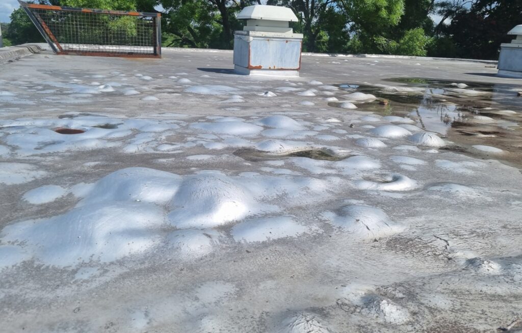 Severely damaged school flat roof with large blisters and water pooling, requiring urgent capital support for repair or replacement.