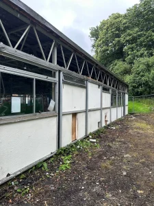 Heathside School - Case Study - Exterior view of an old school building with damaged white panel walls, broken windows, and exposed steel roof trusses, surrounded by overgrown vegetation and debris.