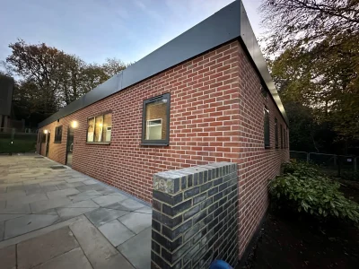 Newly constructed red brick school building with large windows, exterior lighting, and surrounding landscaping at dusk.