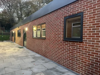 Side view of a modern red brick school building with large windows and a paved walkway, surrounded by trees.