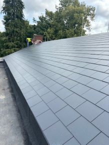 Newly installed pitched slate roof with a worker completing finishing touches near a chimney, surrounded by trees.
