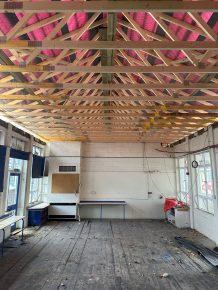 Interior of a classroom under refurbishment with exposed timber roof trusses and visible pink insulation between rafters.