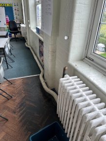 Old classroom radiator with exposed pipework running along the wall beneath windows, highlighting outdated heating infrastructure.