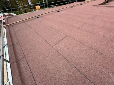 Flat roof covered in newly installed red mineral felt, surrounded by scaffolding for safety during construction.